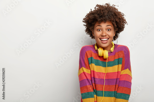 Portrait of pleased millennial Afro girl enjoys spare time  listens music via headphones  looks happily at you  gets pleasant news  wears striped sweater  poses against white studio wall  blank space