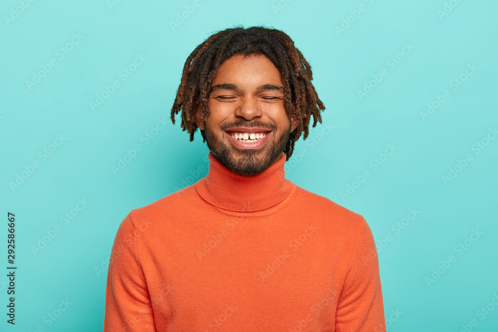 Carefree smiling dark skinned millennial guy has happy facial expression, laughs at something positive, shows white teeth, wears orange poloneck, enjoys spare time, has dreadlocks, poses indoor Carefree smiling dark skinned millennial guy has happy facial expression, laughs at something positive, shows white teeth, wears orange poloneck, enjoys spare time, has dreadlocks, poses indoor