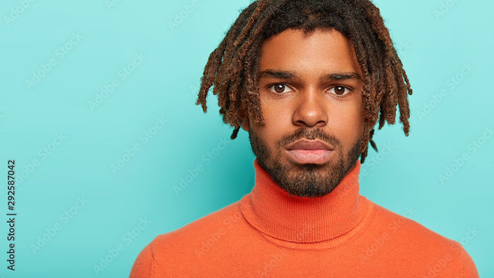 Obraz premium Headshot of serious youngster has dark eyes, healthy groomed skin, has dreadlocks, thick bristle, has self assured gaze at camera, wears comfortable clothes, isolated on blue wall, free space for text