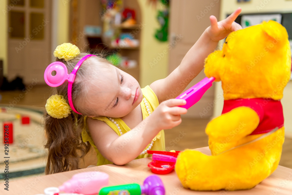 Girl with down syndrom playing doctor with toy bear, rehabilitation center - obrazy, fototapety, plakaty 
