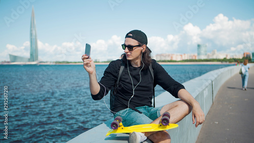 A young man sitting on the embankment with a yellow skateboard on his lap makes a photo on smartphone of the cityscape. © Ulia Koltyrina