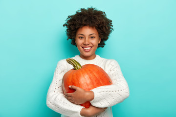 Autumn harvesting, food and Halloween holiday concept. Cheerful curly African American woman embraces ripe pumpkin, smiles pleasantly, dressed in white sweater. Girl with thanksgiving symbol.