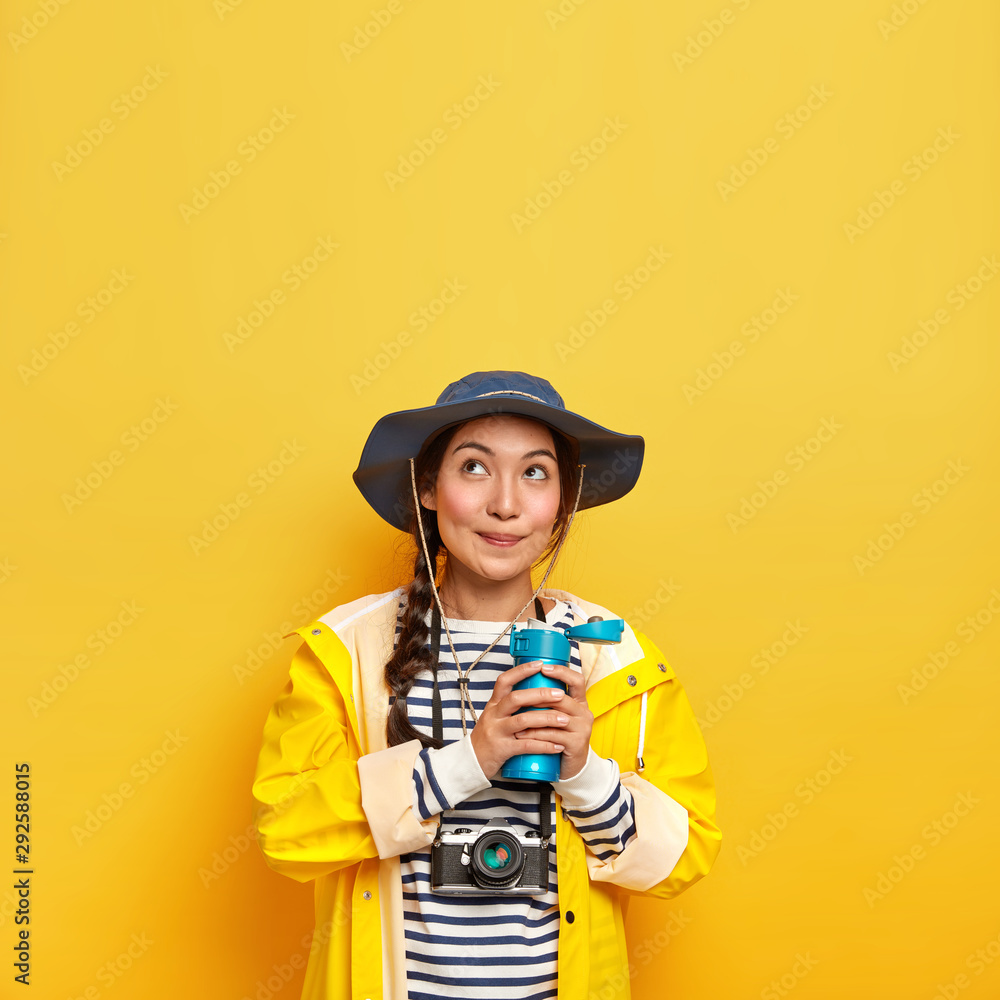 Vertical image of lovely thoughtful girl in stylish headgear, protective raincoat, holds thermos with hot drink, carries retro camera for making photographes, creats content for social media channel Vertical image of lovely thoughtful girl in stylish headgear, protective raincoat, holds thermos with hot drink, carries retro camera for making photographes, creats content for social media channel