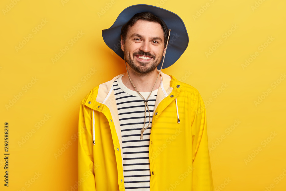 Horizontal shot of pleased Caucasian man wears hat, yellow raincoat, spends leisure time actively, poses indoor, expresses good emotions, has stubble, stands indoor. Happy male model travels alone Horizontal shot of pleased Caucasian man wears hat, yellow raincoat, spends leisure time actively, poses indoor, expresses good emotions, has stubble, stands indoor. Happy male model travels alone