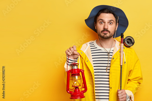Surprised fisherman holds fishing rod and kerosene lamp, has overnight fishing trip, wears hat and raincoat, poses over yellow background, copy space for your advertising area. Angling concept photo