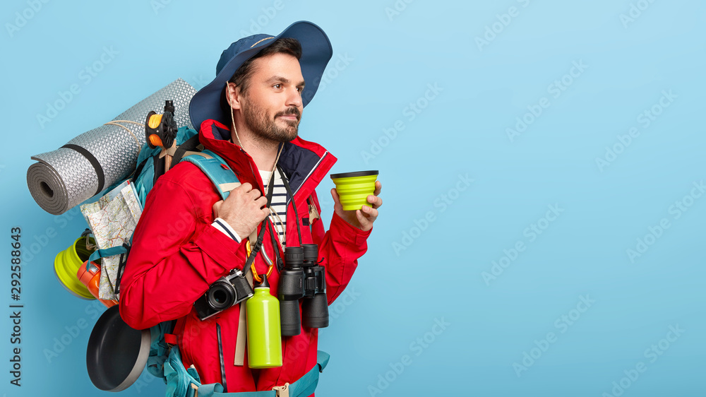 Young thoughtful man in trendy hat and red jacket, drinks coffee, walks in forest with backpack, poses against blue background, thinks about new destination, makes stop during traveling or vacation Young thoughtful man in trendy hat and red jacket, drinks coffee, walks in forest with backpack, poses against blue background, thinks about new destination, makes stop during traveling or vacation