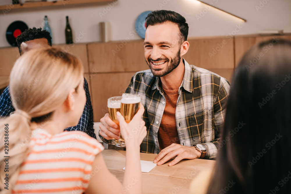 smiling man clinking glasses of light beer with young woman while sitting in pub with multicultural friends