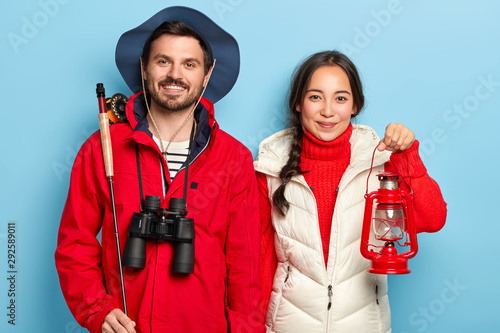 Pleased mixed race couple have night fishing together, woman holds kerosene lamp, man stands with fishing rod and binoculars, enjoy camping journey, stand next to each other against blue background photo