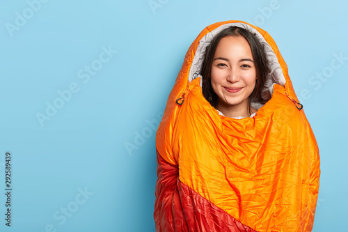 Pleased young Asain brunette woman wrapped in warm orange sleeping bag, spends free time actively, being avtive camper, stands against blue background, blank space for your advertisement or text photo