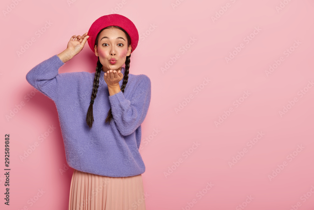 Adult korean woman keeps lips rounded, sends air kiss, blows mwah, keeps one hand on red beret, wears makeup, oversized purple sweater, expresses romantic passionate feelings, models indoor. - obrazy, fototapety, plakaty 