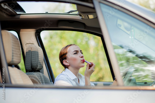 Concept of danger driving. Young woman driver red haired teenage girl painting her lips doing applying make up while driving the car © volody10