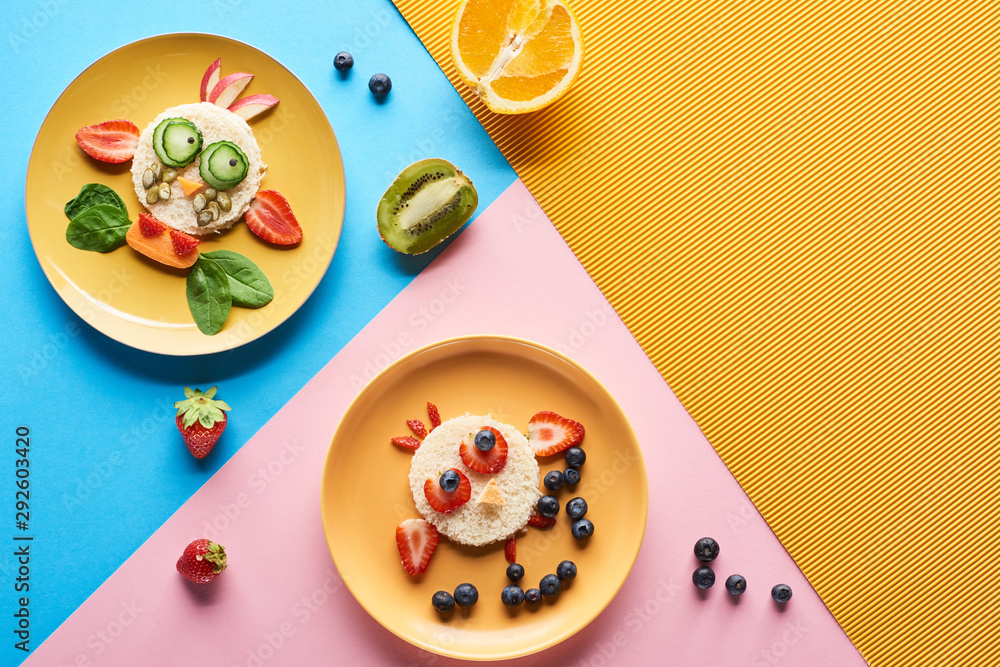 Fototapeta widok z góry na talerze z fantazyjnymi zwierzętami z jedzenia na śniadanie dla dzieci na niebieskim, żółtym i różowym tle
