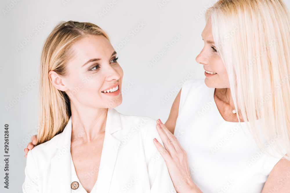 smiling blonde mature mother looking at young daughter in total white clothes isolated on grey smiling blonde mature mother looking at young daughter in total white clothes isolated on grey
