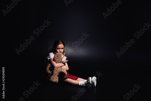 Fototapeta Naklejka Na Ścianę i Meble -  lonely, scared kid sitting on floor and hugging teddy bear on black background