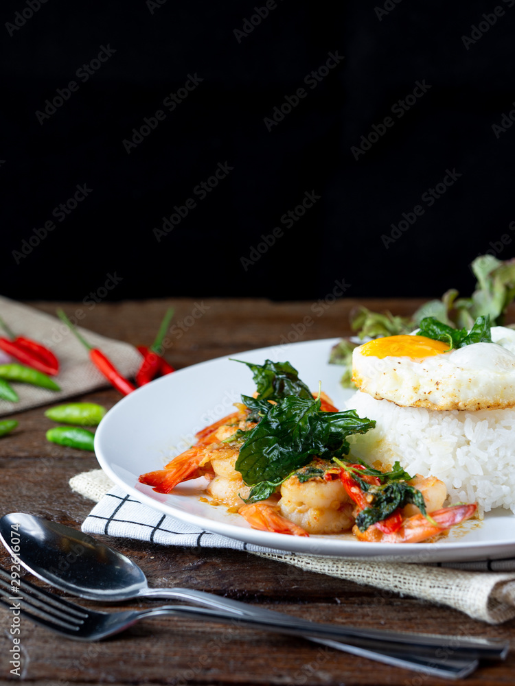 Spicy fried shrimp with basil and chili, serve with rice and egg in white ceramic plate on wood table, Thai style food.