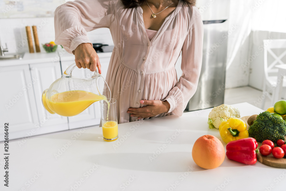 cropped view of pregnant woman pouring orange juice in glass near vegetables