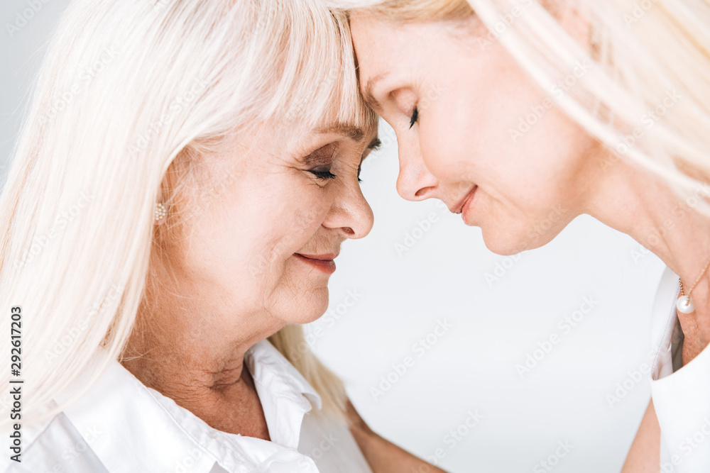 blonde mature daughter and senior mother standing face to face with closed eyes isolated on grey blonde mature daughter and senior mother standing face to face with closed eyes isolated on grey
