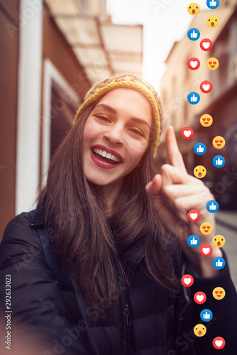 A beautiful young woman blogger, vlogger or influencer is receiving emoji and emoticon reactions in her mobile smart phone device while making a post, sharing or video logging on social media