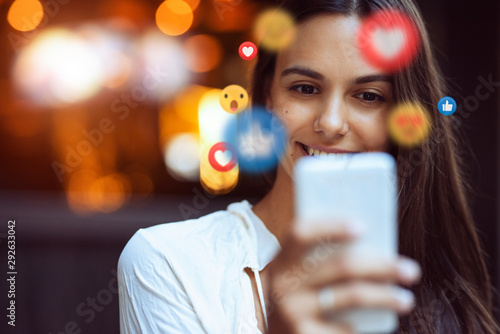 A beautiful young woman blogger, vlogger or influencer is receiving emoji and emoticon reactions in her mobile smart phone device while making a post, sharing or video logging on social media photo