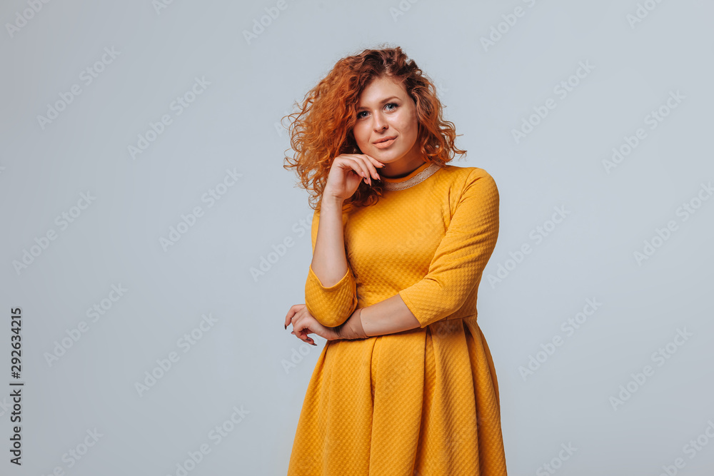 Fototapeta premium A red haired girl in a yellow dress on a light gray background smiles and touches her face