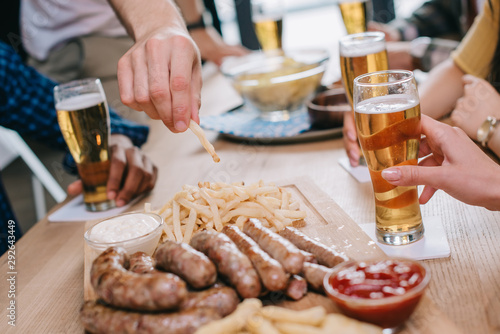 cropped view of man taking french fries and fried sausages on wooden tray in pub