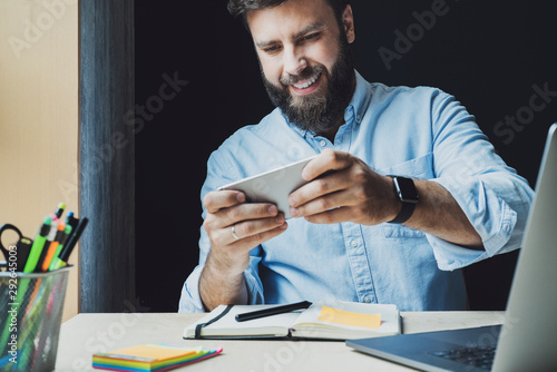 Man in blue shirt sitting in front of laptop, using smartphone. Smiling guy is playing game on smartphone. Online marketing.