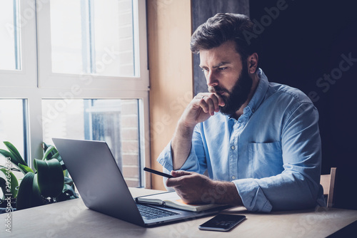 Professional sitting in office in front of laptop. Developer thinking on solutions for work. Home-based student getting distant education. Young serious bearded man in blue shirt working on desktop.