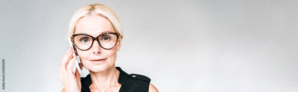 panoramic shot of mature businesswoman in black outfit and glasses talking on smartphone isolated on grey panoramic shot of mature businesswoman in black outfit and glasses talking on smartphone isolated on grey