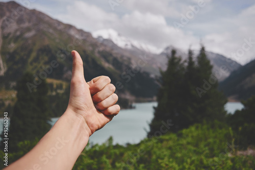 Thumb up on the background of Mountain Lake Bao in Almaty. Beautiful nature, summer green landscape of sunny Kazakhstan