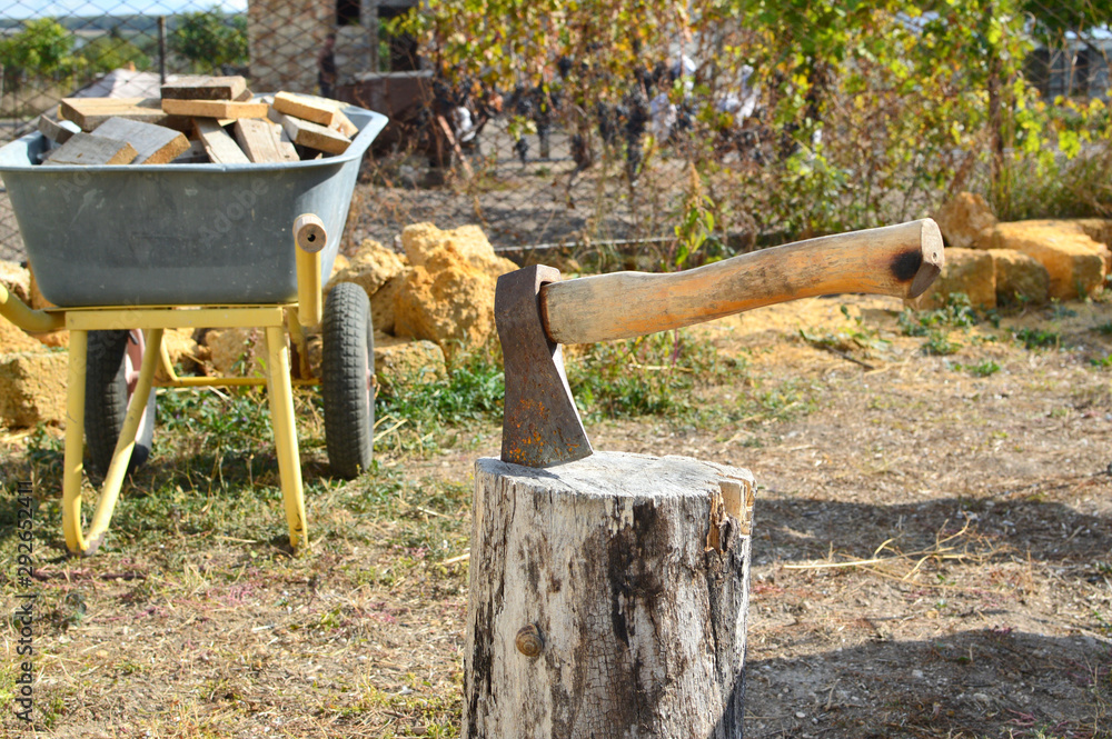 Ax on a wooden log. in the background a wheelbarrow with a bunch of wooden firewood. Ax on a wooden log. in the background a wheelbarrow with a bunch of wooden firewood.