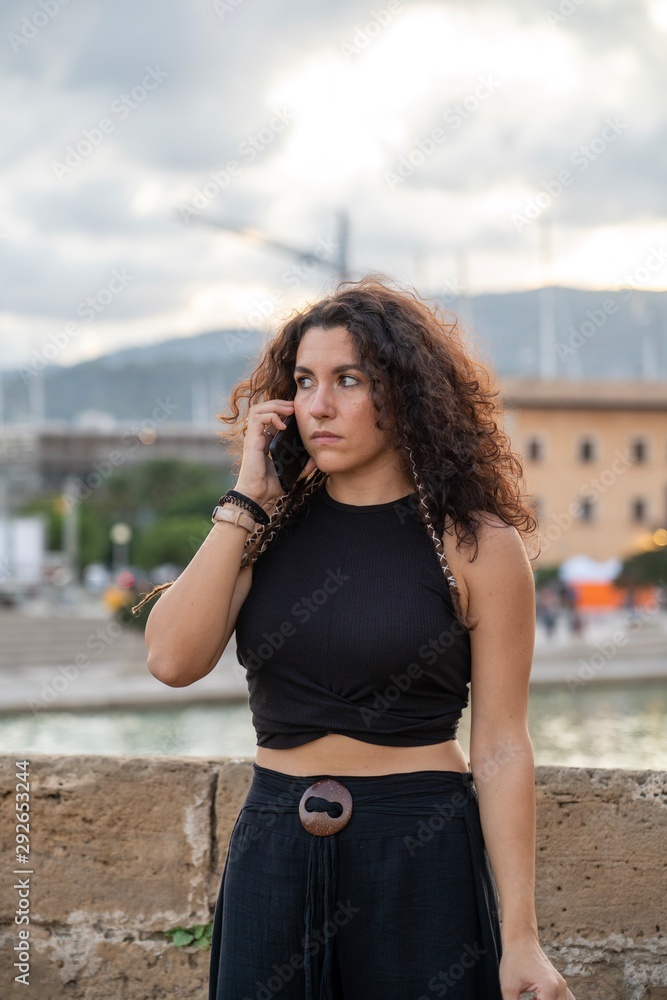 Young caucasian brunette woman with curly hair and dreadlocks standing serious while talking on the phone