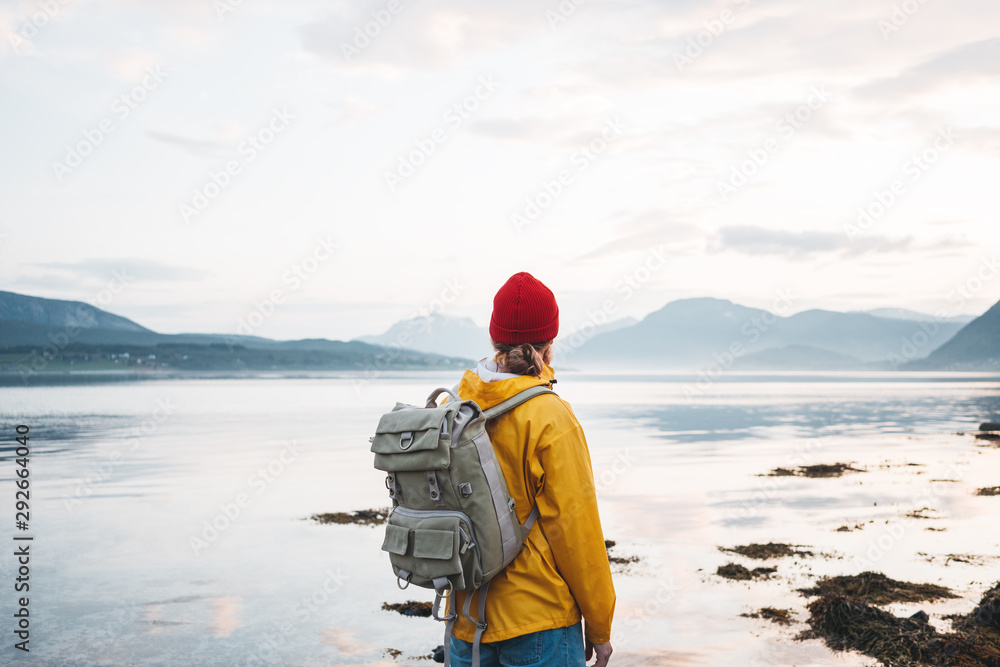 Hipster traveler wearing backpack and yellow raincoat looking away at mountain and fjord. Alone man traveling at scandinavian authentic landscape