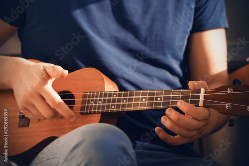 A man in a dark blue t-shirt and blue jeans plays a cheerful tune on a ukulele, sitting in the sunlight.