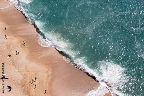 Drone view of people on the yellow sand beach facing the Beautiful crushing waves of Atlantic ocean in Nazare, Portugal