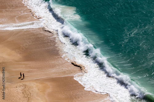 Drone view of people on the yellow sand beach facing the Beautiful crushing waves of Atlantic ocean in Nazare, Portugal 