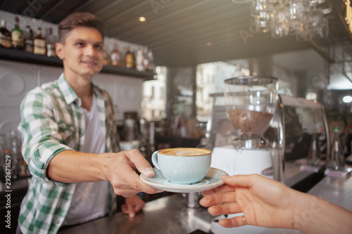Cropped shot of a cheerful male barista giving a cup of coffee to unrecognizable customer. Handsome man working at his coffee shop, selective focus on a cup of coffee