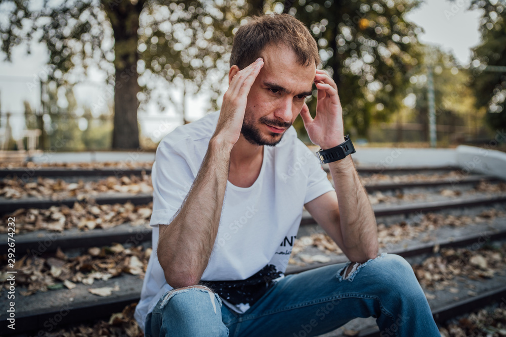 A handsome man in depression. Not shaved hipster sits on the steps in the autumn Park holding his head. Concept: the headache and grief A handsome man in depression. Not shaved hipster sits on the steps in the autumn Park holding his head. Concept: the headache and grief