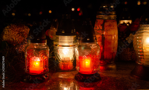 Candles in elegant glass lanterns on a tombstone - in the background flower bouquet - all souls night decoration in the dark - european christian cemetery