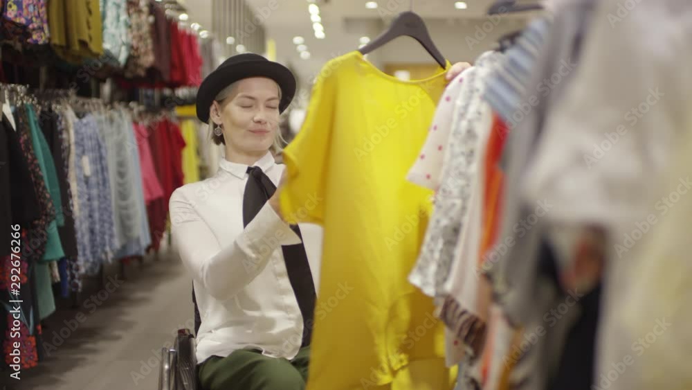 Tracking of happy beautiful woman in wheelchair smiling and taking photo of yellow blouse in clothing store, then posing it on social media or sending to friend Tracking of happy beautiful woman in wheelchair smiling and taking photo of yellow blouse in clothing store, then posing it on social media or sending to friend