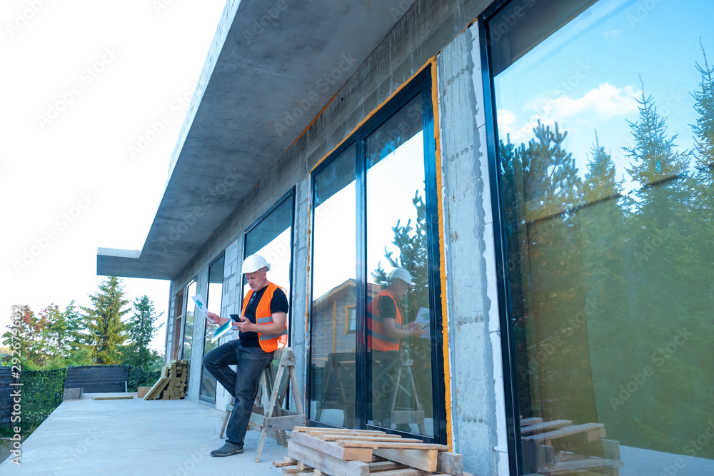 The Builder stands next to the building under construction. Panoramic Windows of the house. The study of construction documentation for the building. Engineer builder is studying blueprints. The Builder stands next to the building under construction. Panoramic Windows of the house. The study of construction documentation for the building. Engineer builder is studying blueprints.