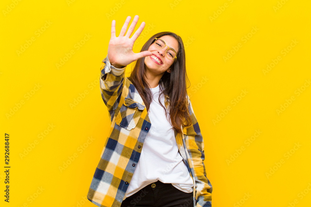 young  pretty woman smiling and looking friendly, showing number five or fifth with hand forward, counting down against orange wall - obrazy, fototapety, plakaty 