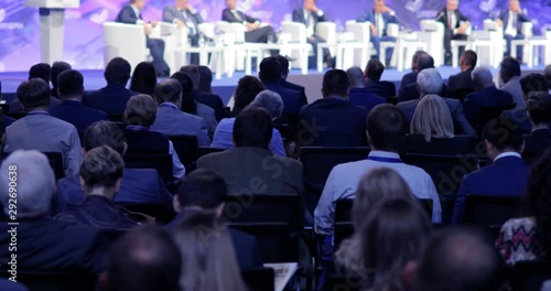 Politicians or scientists speak from the stage, people in audience sitting back to the camera. Spectators and journalists at scientific or political press conference, briefing, view from auditorium.