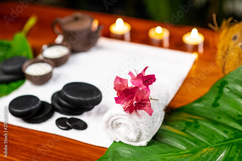 A terry towel on which there is a clay teapot and cups for drinks with milk, stones for stone therapy, lighted small candles, a mangolia flower and a twisted terry towel on a wooden table