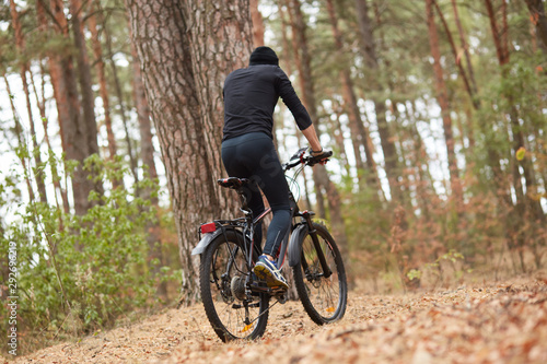 Handsome young man biking in countryside, guy wearing black sportwear and cap posing backwards around trees in wood, spending spare time in open air, enjoys beautiful nature. Sport and fitness concept