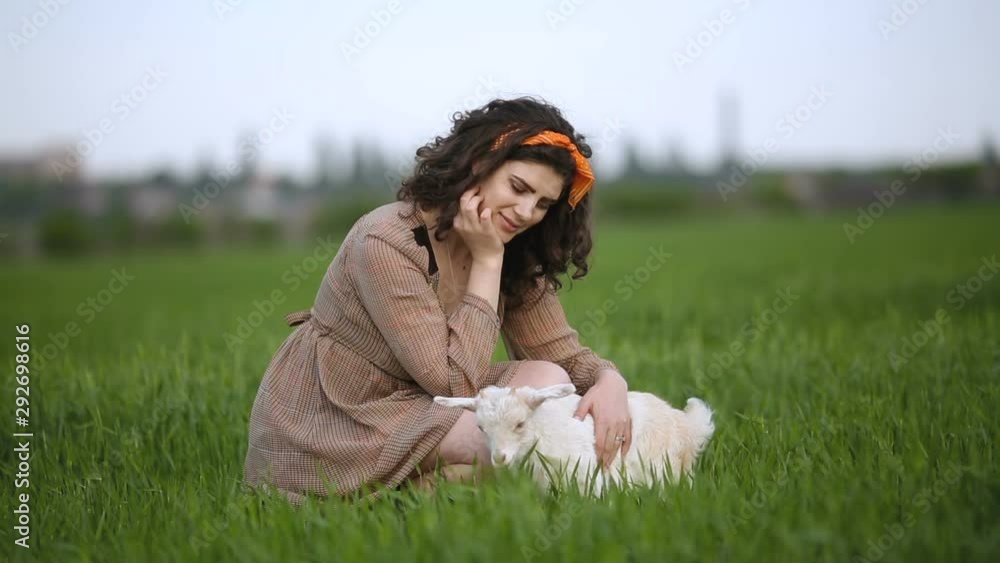 Pretty village girl sitting in the field with little white goat. Beautiful plus-size model posing. Friendship of people and animals. Vegetarian concept. Farmer on small farm. Pretty village girl sitting in the field with little white goat. Beautiful plus-size model posing. Friendship of people and animals. Vegetarian concept. Farmer on small farm.