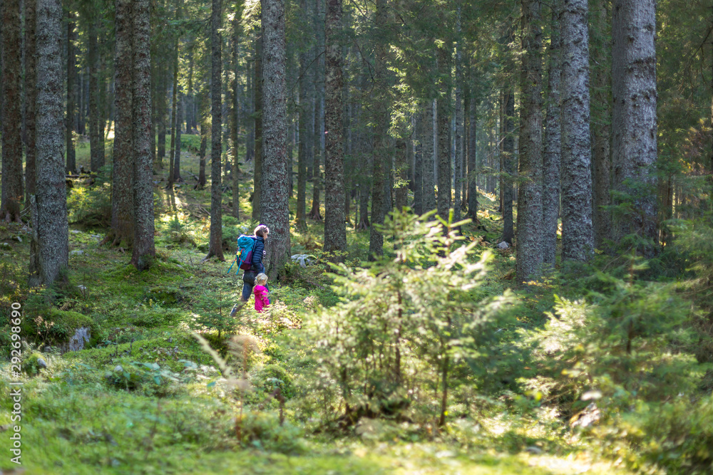 Obraz premium Hiker mother with backpack and daughter walking on path and exploring summer spruce forest. Enjoying the pristine nature.