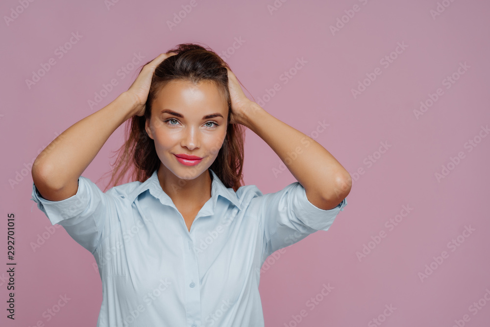 Isolated shot of attractive young woman keeps both hands on hair, looks pleasantly at camera, has natural beauty, wears light blue shirt, poses against purple background, empty space for your text