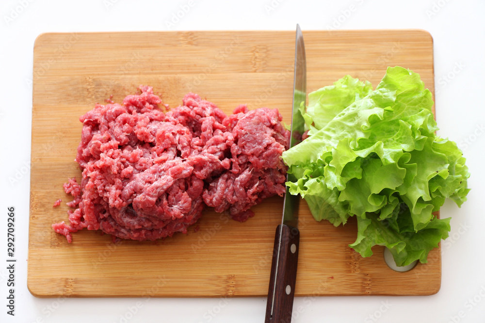 Plant-based meat concept. On a wooden cutting board are green leaves of lettuce salad and red ground beef, separated by a knife with a wooden handle. White background.