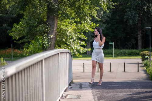 Businesswoman is walking through the park and talking on the phone. Woman in white top and skirt on the phone on a sunny day in the park with business customers. Smiling woman on the phone © JürgenBauerPictures