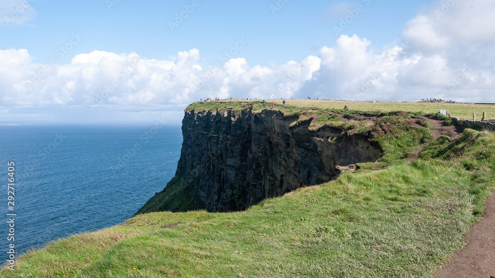 The famous Cliffs of Moher in County Clare, Ireland on a sunny summer day with tourists walking along the top of the cliff.